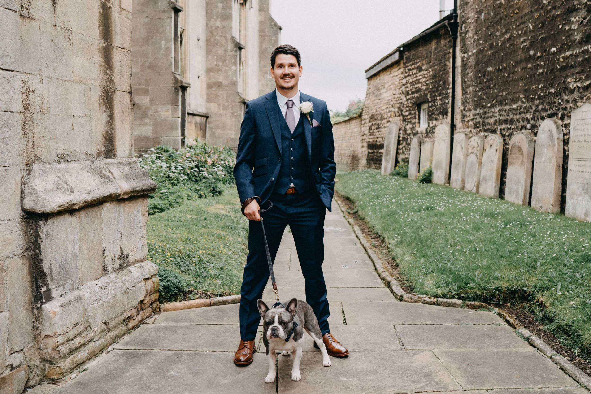 A groom in a navy suit stands with a small gray and white dog on a path between stone church walls.