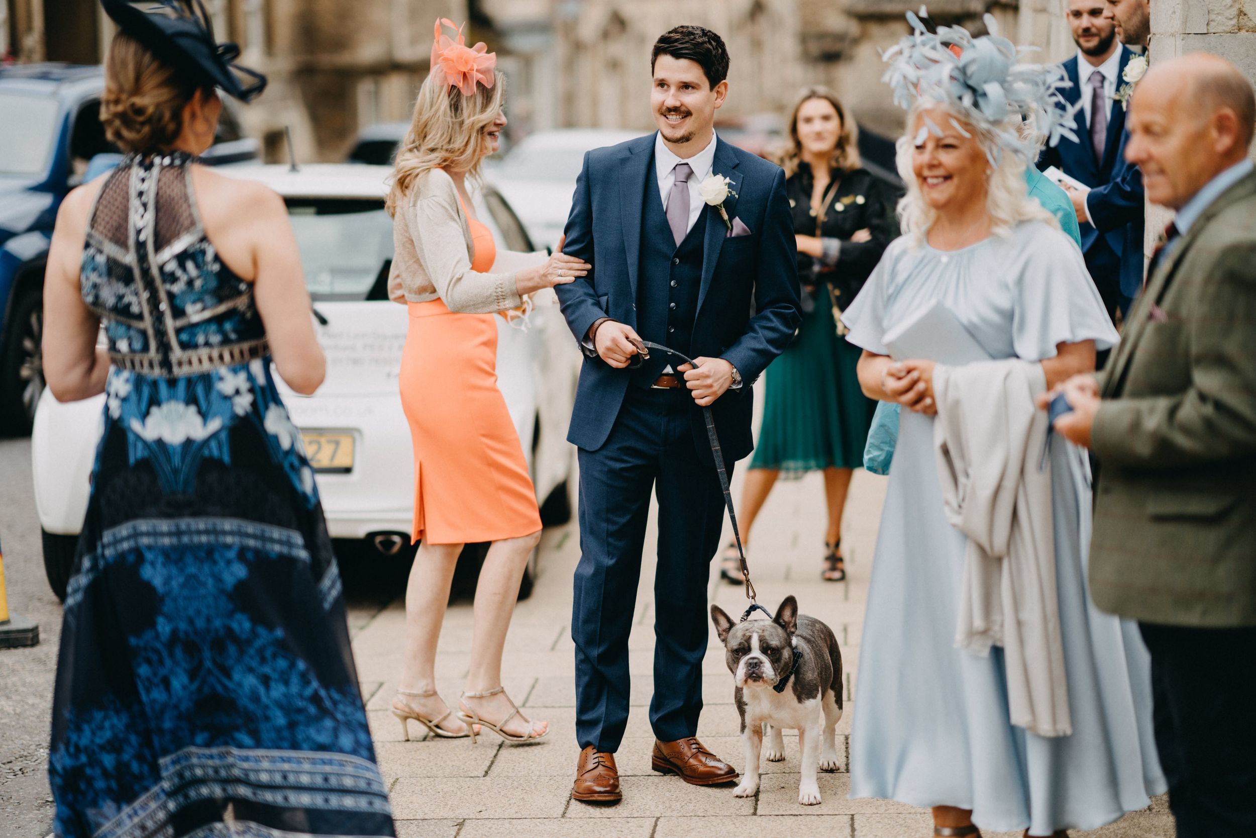 A dog stands among elegantly dressed guests at an outdoor social gathering on a sunny day.