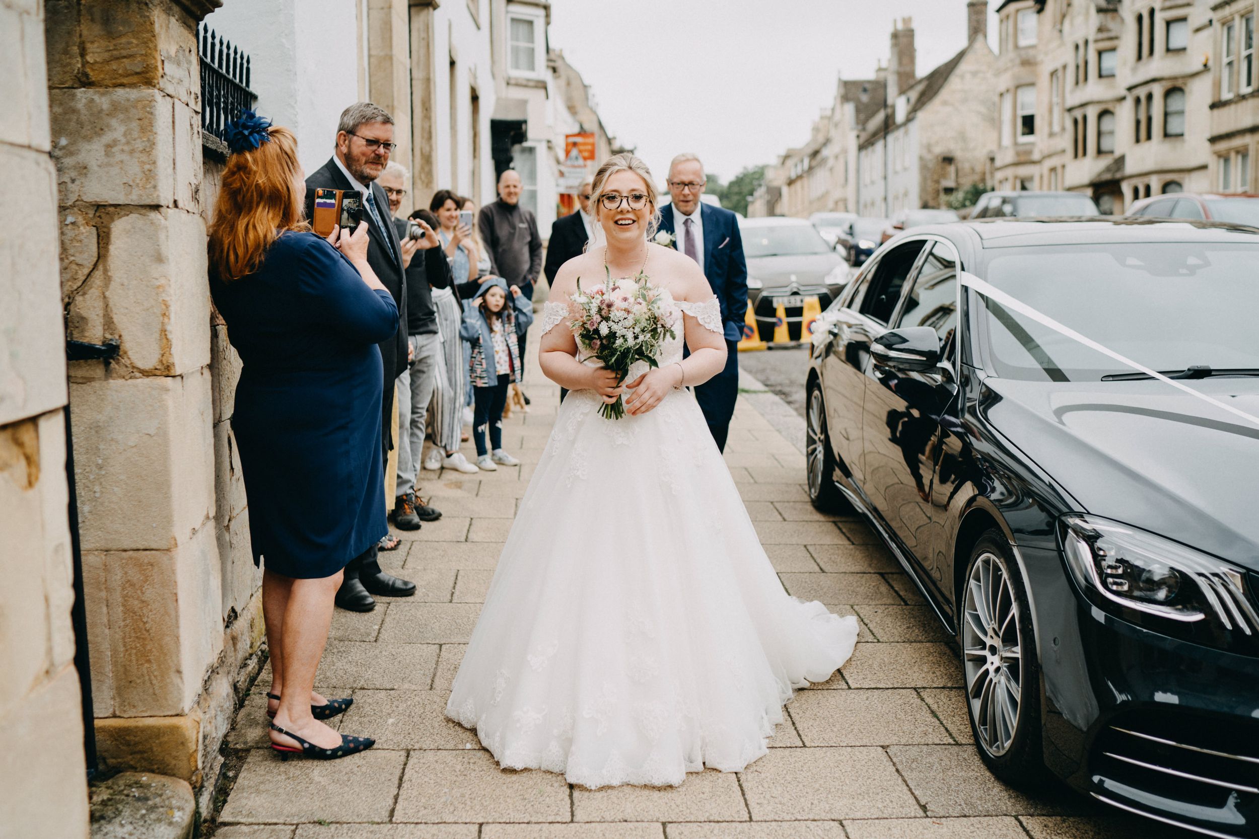 A joyful scene outside with onlookers waving at a person in an elegant white wedding gown next to a black luxury car.