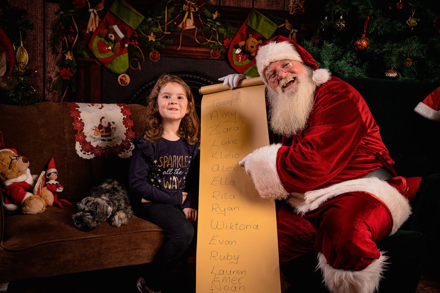 Santa Claus reviews a long scrolled list in a festively decorated indoor Christmas setting.