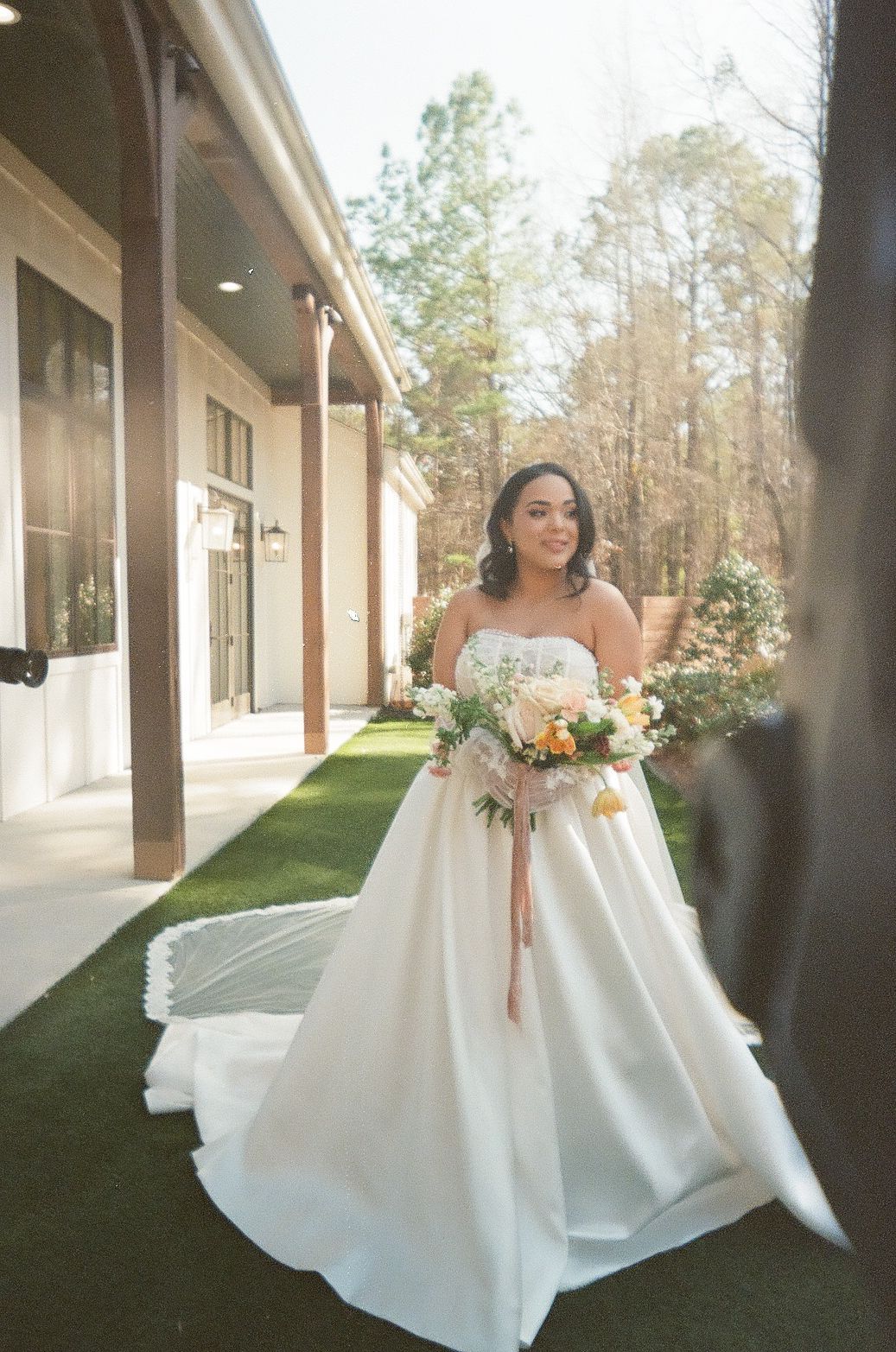 A bride in an elegant white strapless ball gown holds a romantic bouquet outside a modern venue.