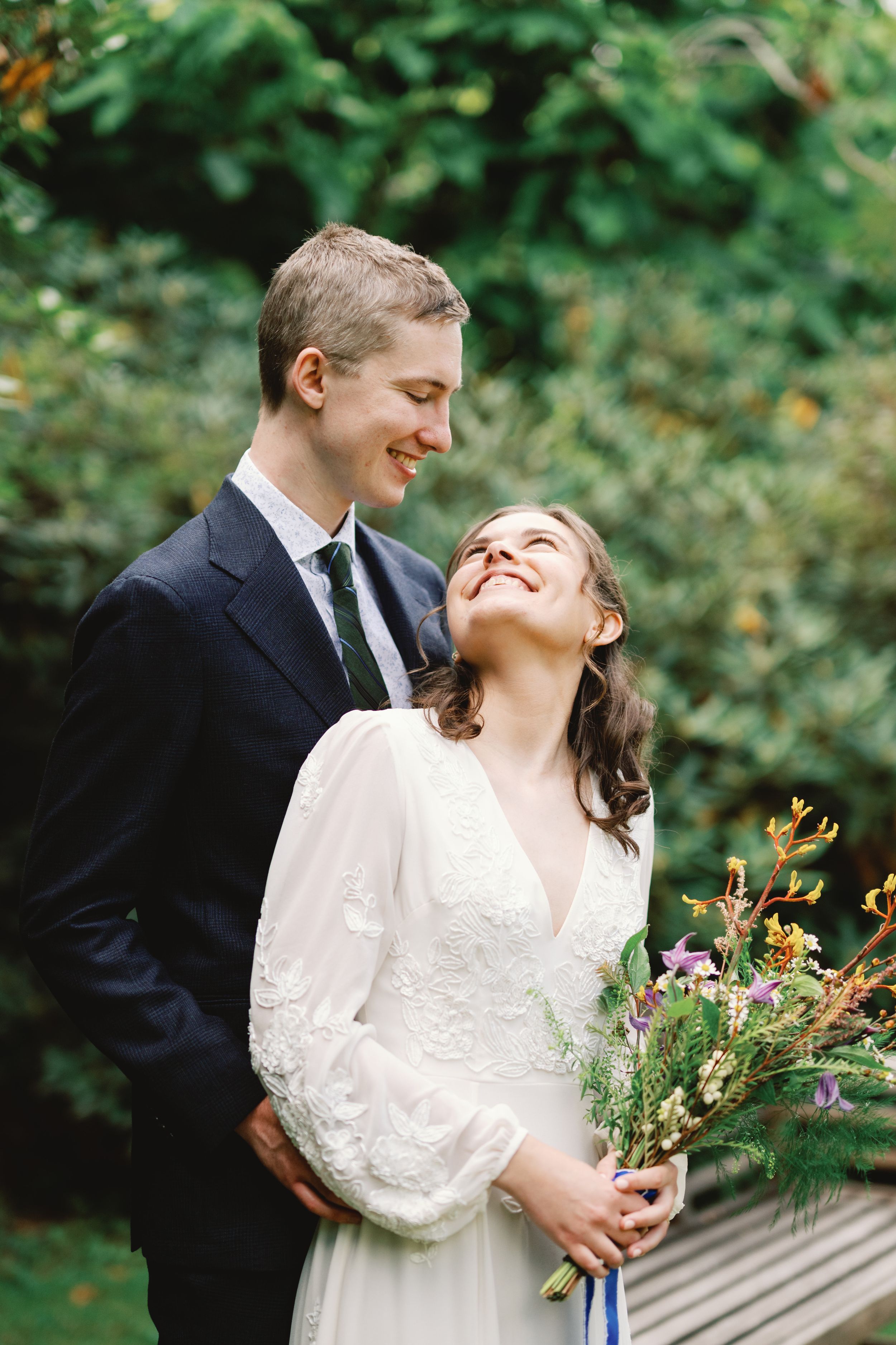 A joyful couple shares a candid moment outdoors during their wedding, with the bride holding a wildflower bouquet.