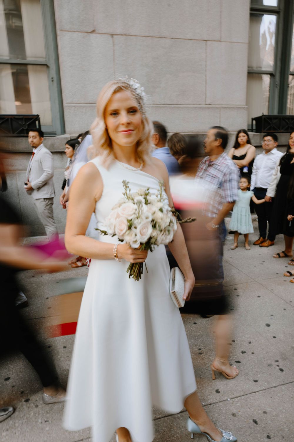 A bride in a white knee-length dress holds a beautiful bouquet of white flowers outside a building with guests in background.