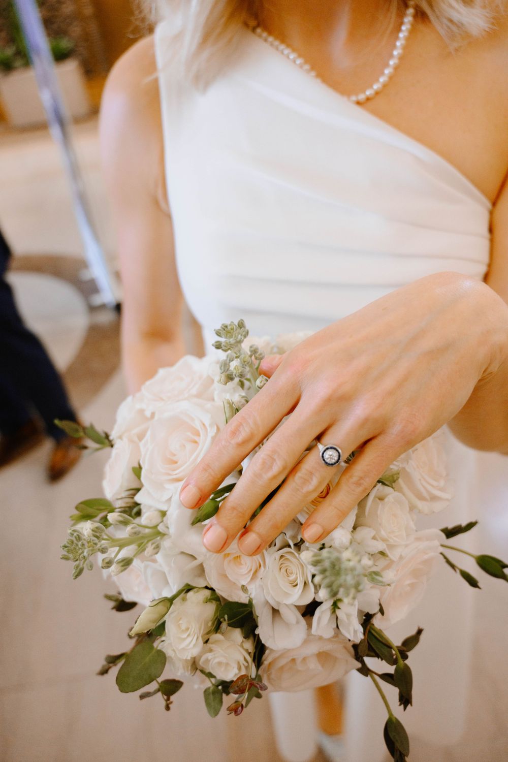A delicate bridal bouquet of blush pink roses and greenery held by someone wearing a one-shoulder white dress and pearl necklace.