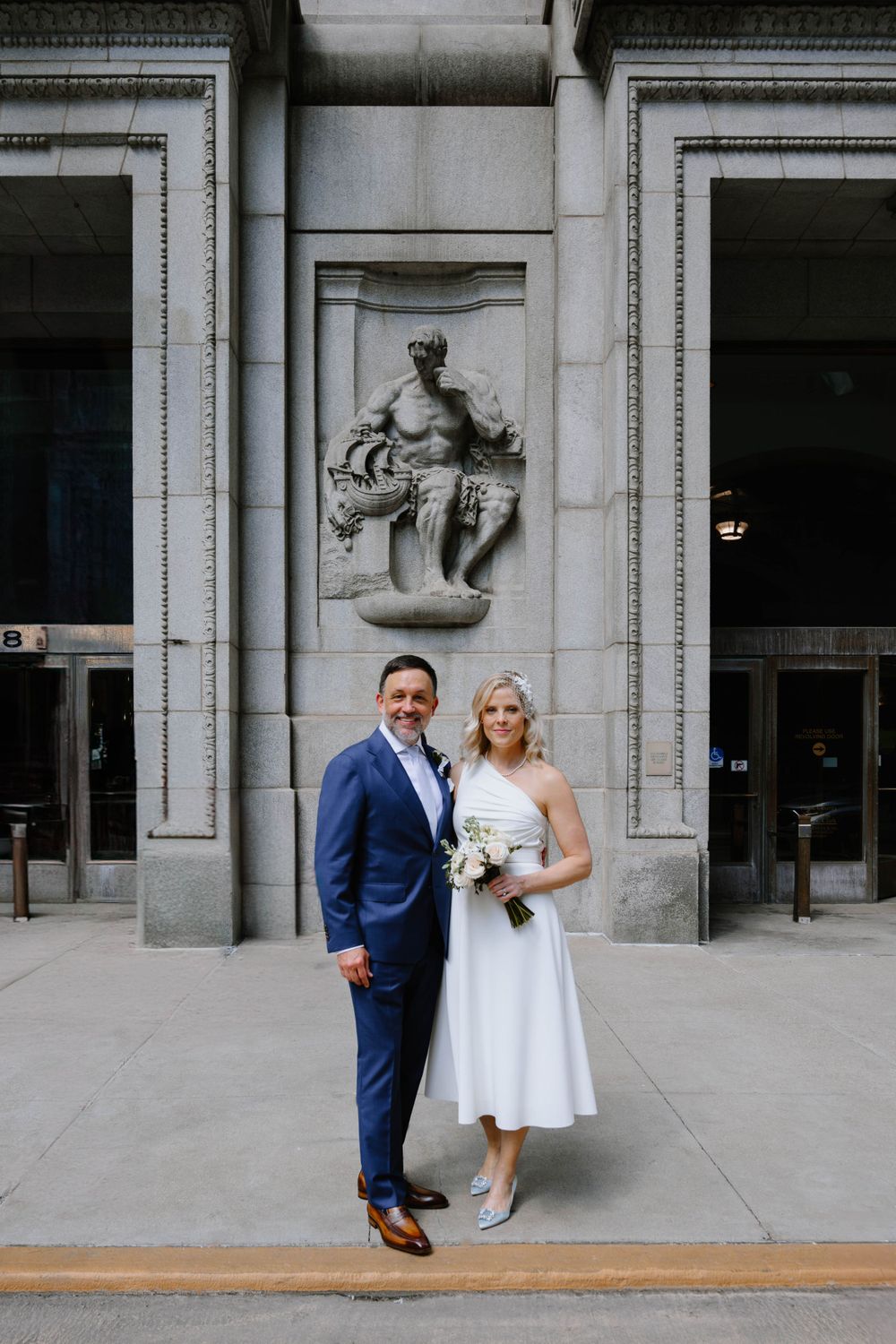 A couple posing in formal attire against a stone building with architectural details and a classical statue.