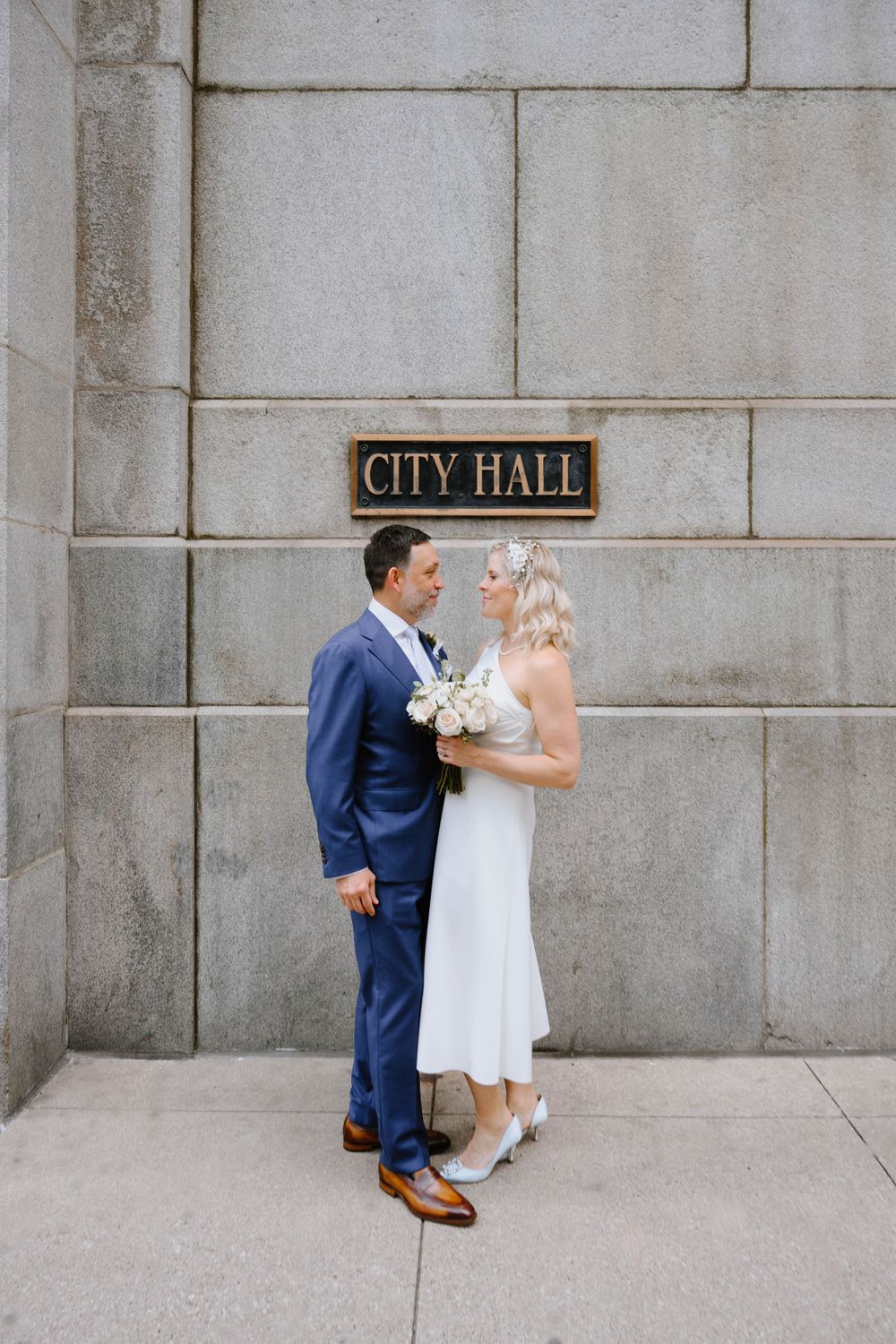 A couple poses for a wedding photo beneath a City Hall sign on a concrete building exterior.