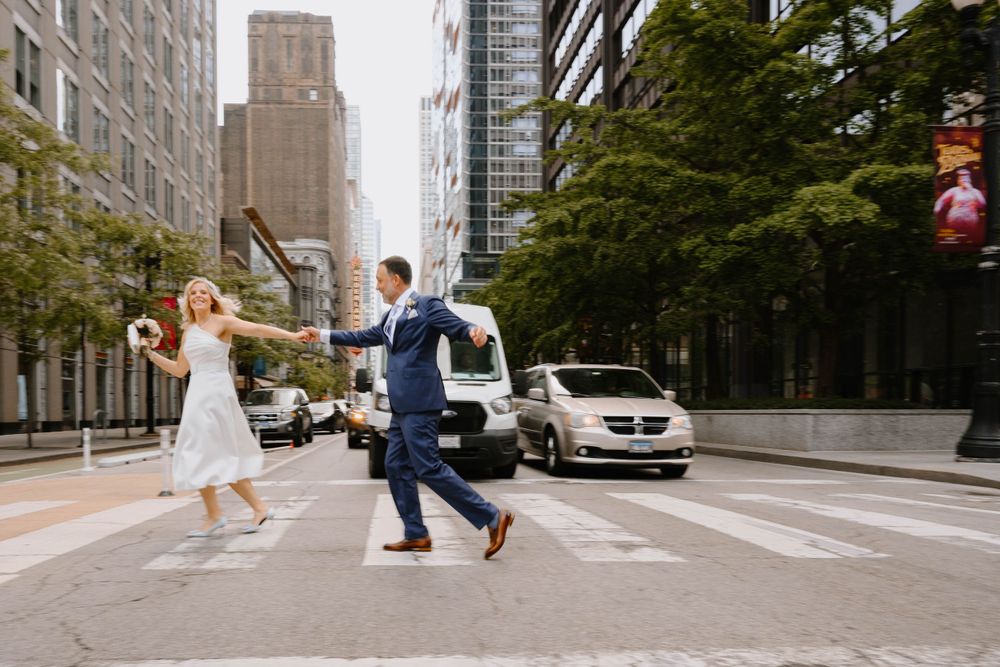 A playful wedding couple runs across a city street with tall buildings in the background.