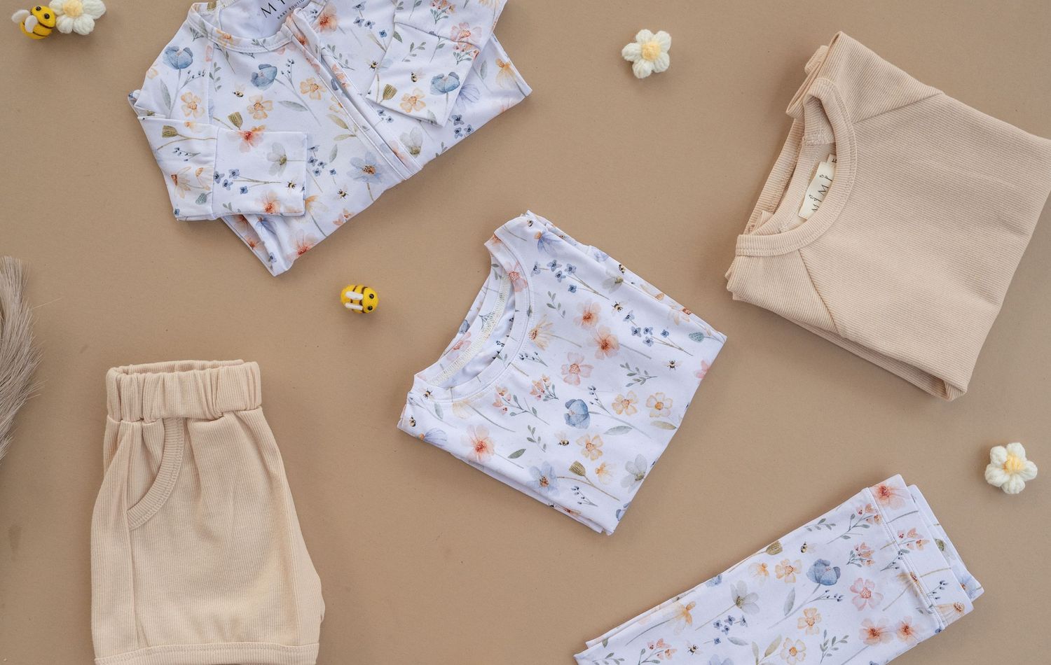 Flat lay of baby clothes including patterned onesies and beige sets on brown background.