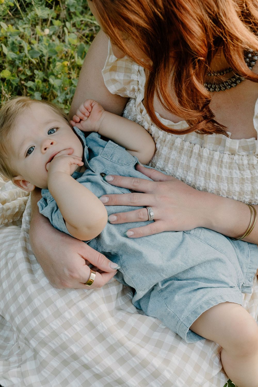 Baby in blue romper lying on checkered blanket outdoors in summer setting.
