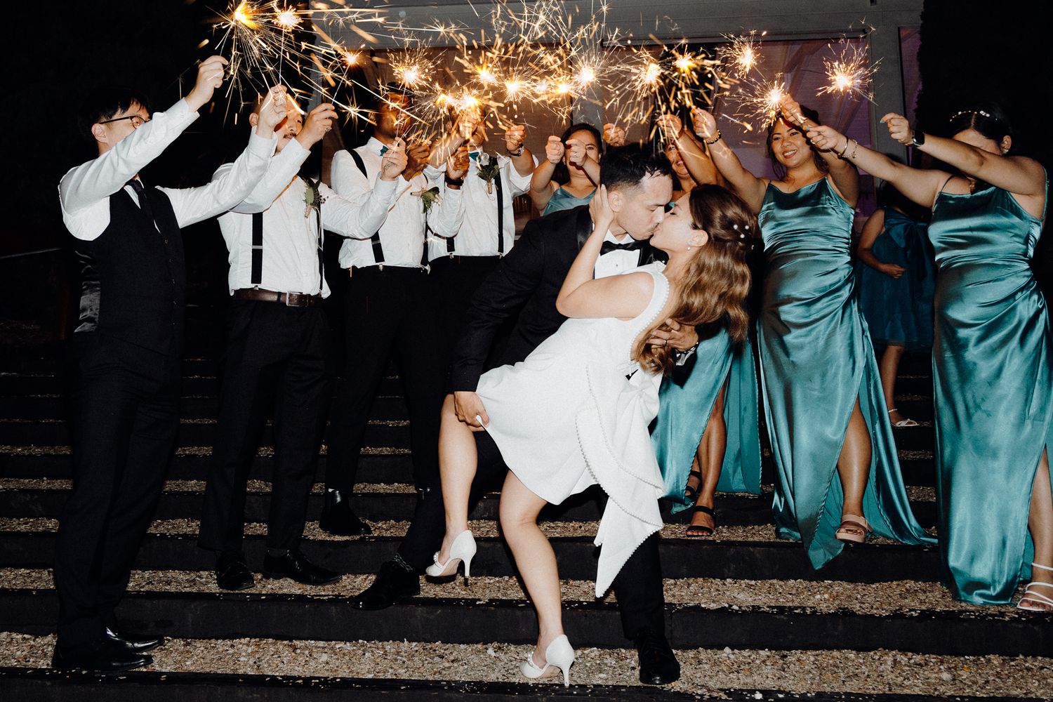 Wedding party wearing teal dresses and black suits pose playfully with sparklers captured by Apertured Media Auckland Wedding Photographer.