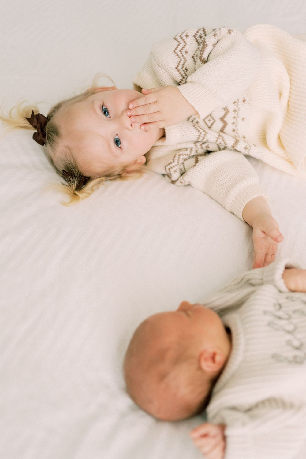 Siblings laying on white bedding wearing matching cream-colored sweaters.
