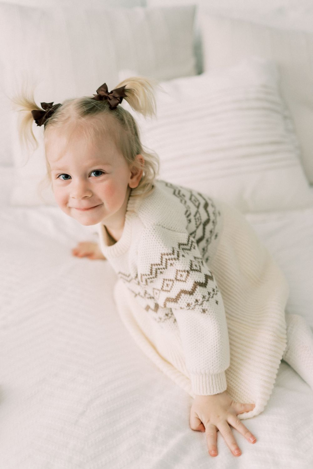 A toddler in a white Fair Isle sweater lying on white bedding.