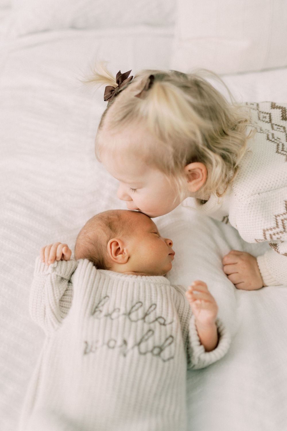 A toddler in a patterned sweater gently kisses a newborn baby on white bedding.