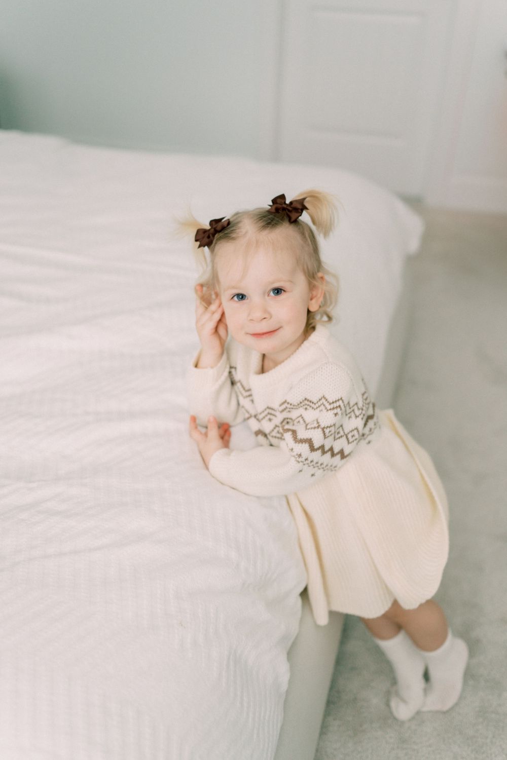 A small child in a cream-colored sweater with bow hair clips sits on white bedding.