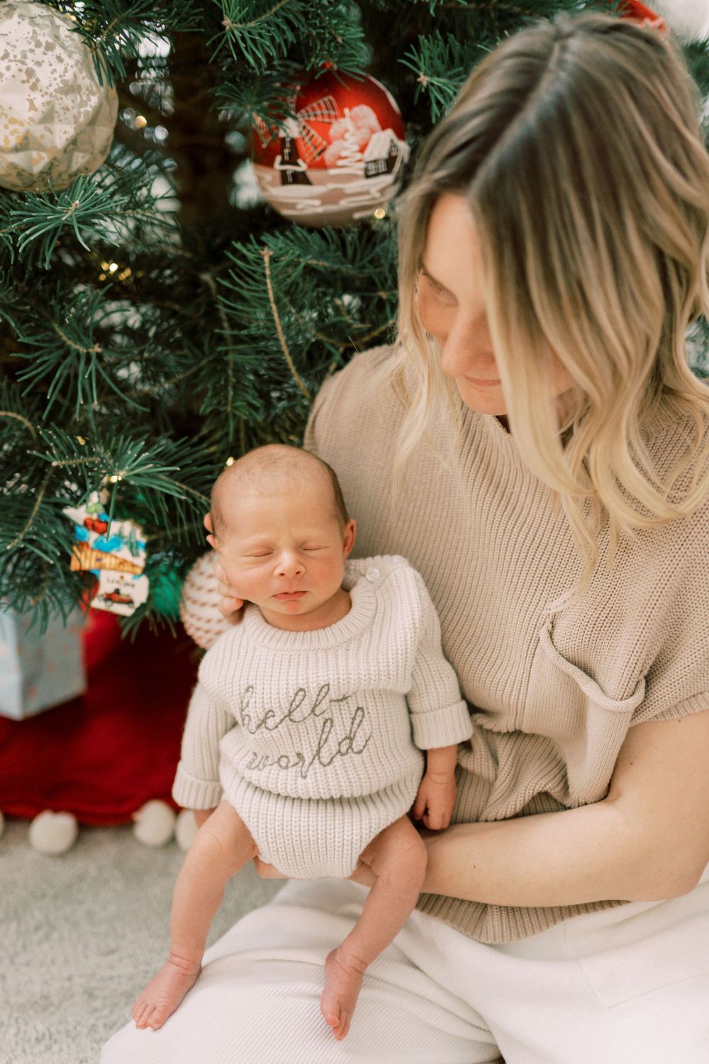 Holiday scene with infant in hello world sweater next to decorated Christmas tree and ornaments.