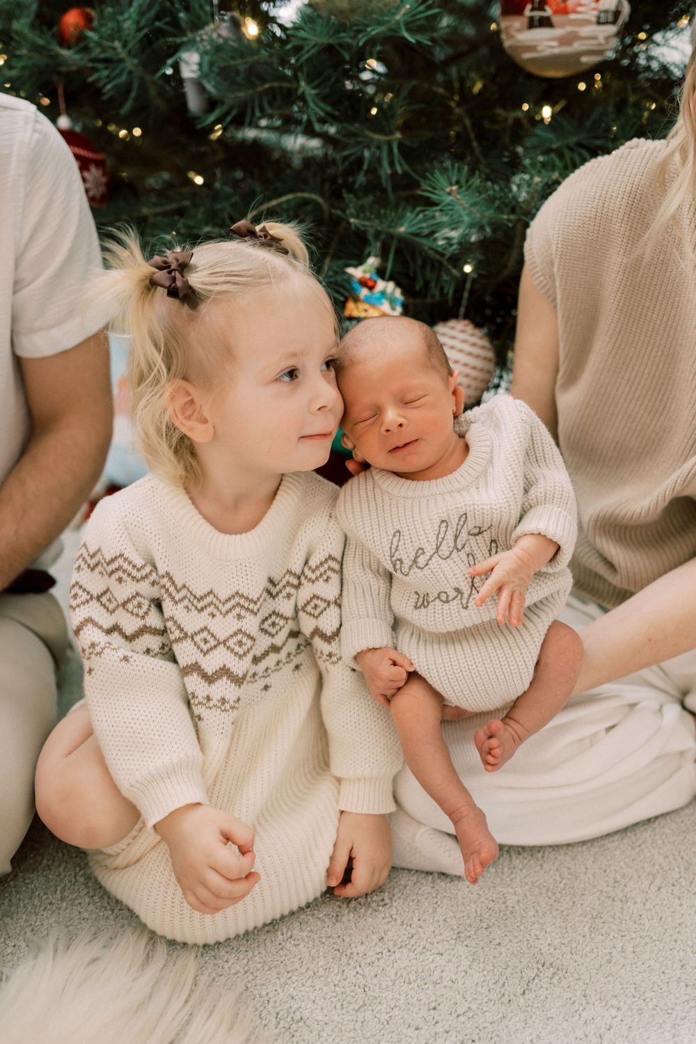 Two children in matching cream knit sweaters sitting by Christmas tree with festive decorations.