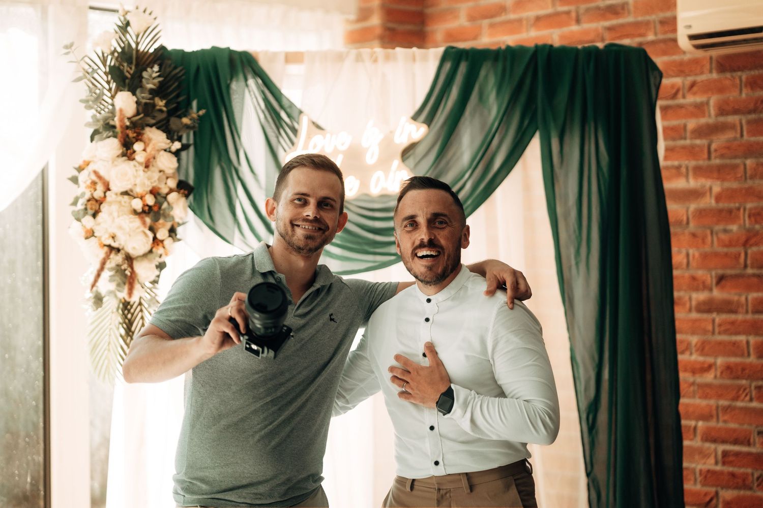 Two friends share a happy moment together in front of emerald green drapes and floral decorations at an event.
