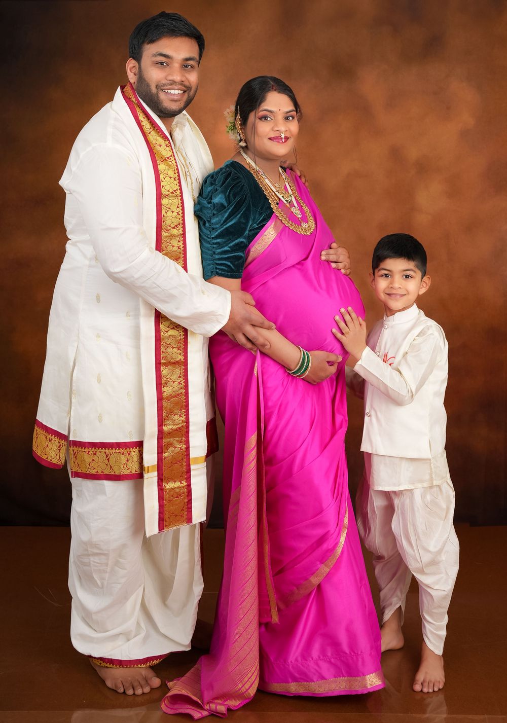 South Asian family maternity portrait in traditional formal attire against brown backdrop.