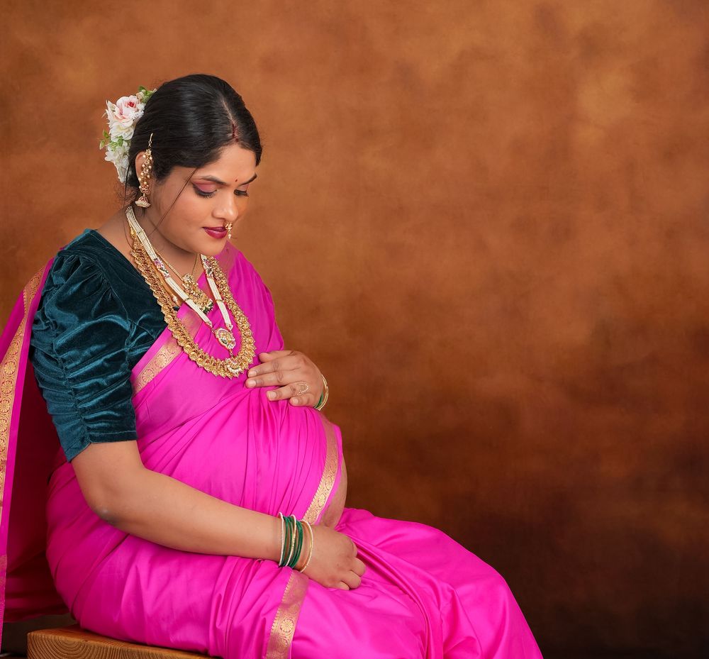 Maternity portrait of woman in bright pink saree and teal blouse against brown backdrop.