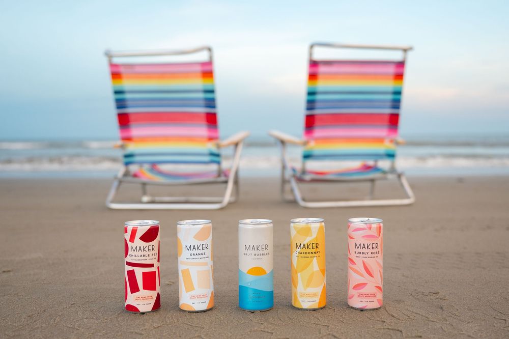 Colorful beach chairs and craft cocktail cans arranged on sandy beach at sunset with ocean waves in background.