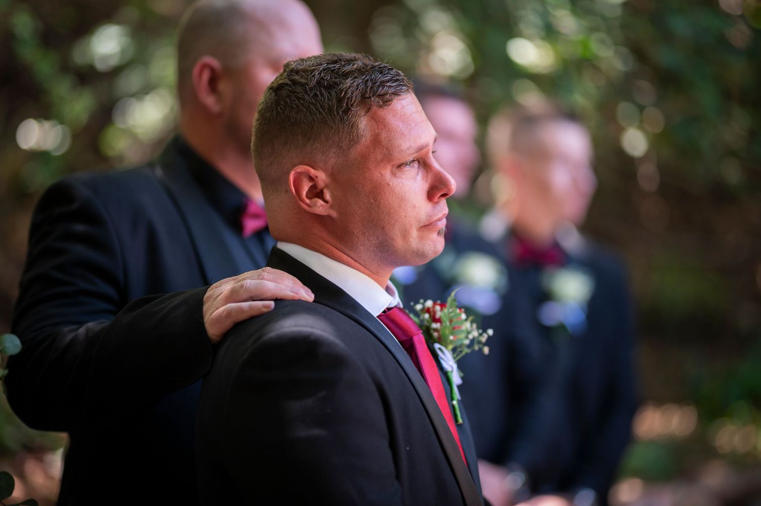 A groom at an outdoor wedding ceremony wearing a black suit with red tie and floral boutonniere.