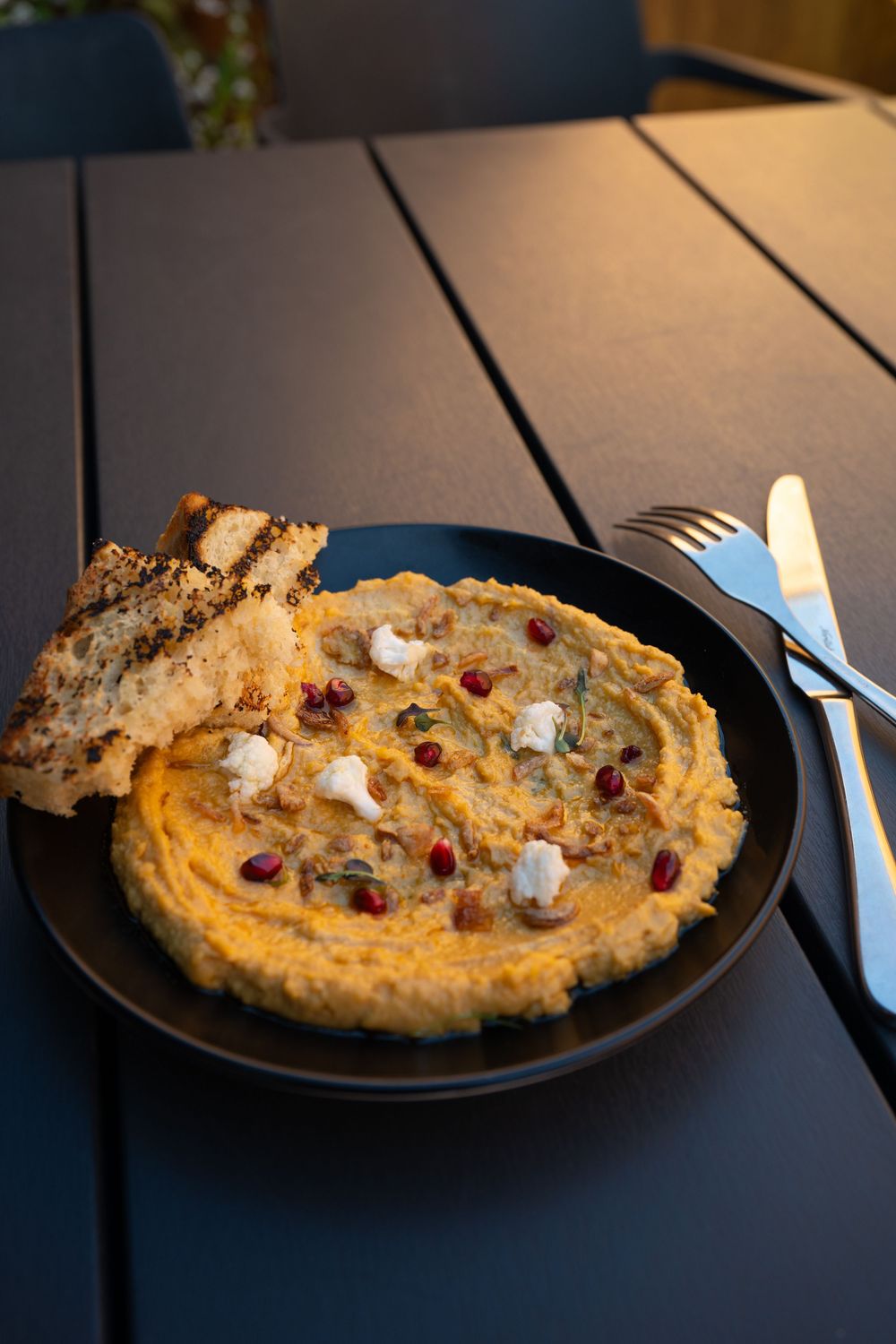Creamy hummus served on a black plate topped with pomegranate seeds, feta cheese and a side of pita bread.