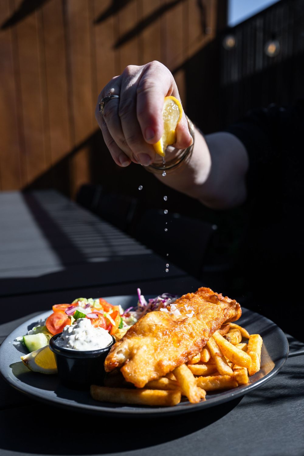 Oil being dripped onto crispy golden fish and chips with tartar sauce and lemon wedges on a black plate.