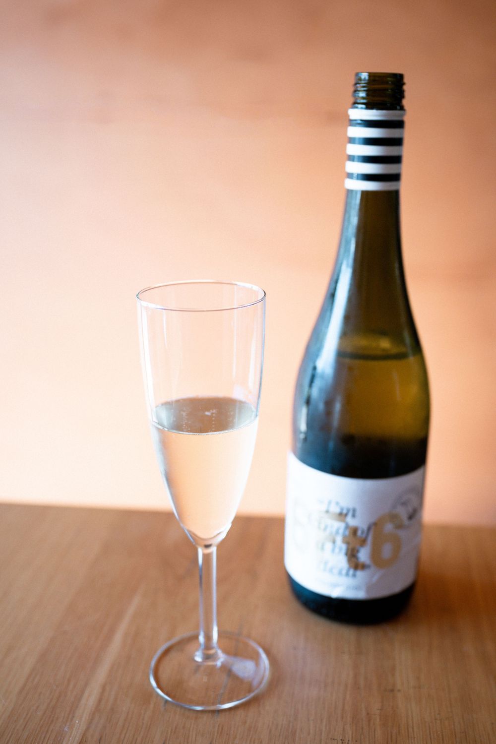 A glass of sparkling wine sits beside an elegant wine bottle on a wooden table with warm lighting.