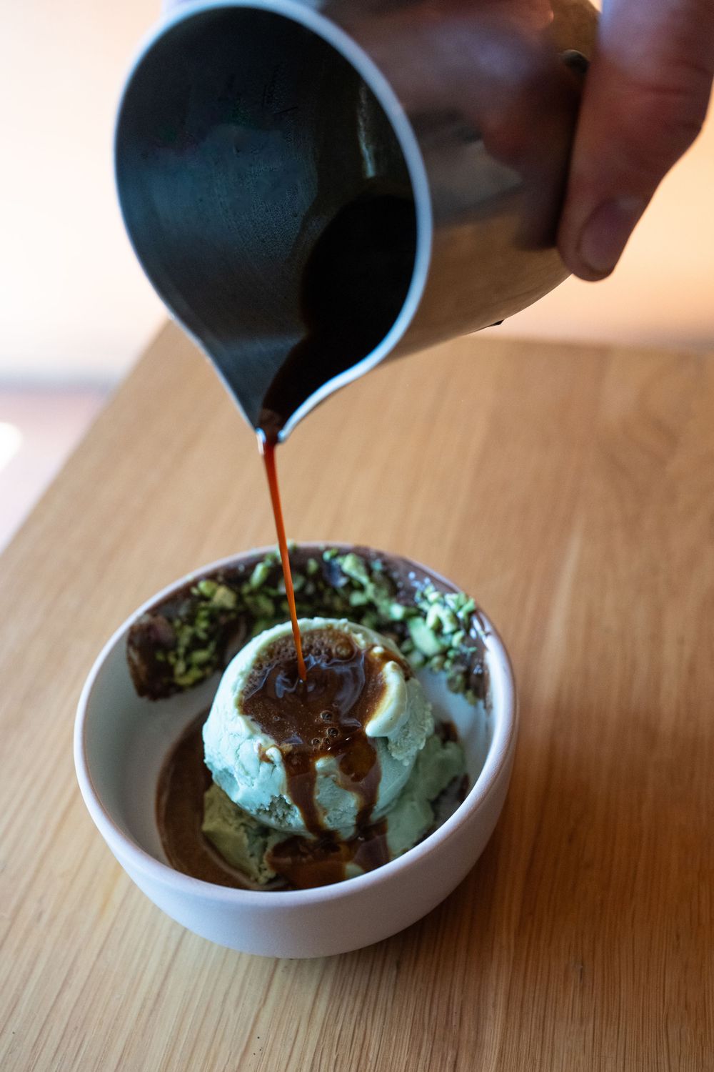 Hot chocolate sauce being poured over mint ice cream in a white ceramic bowl on a wooden surface.