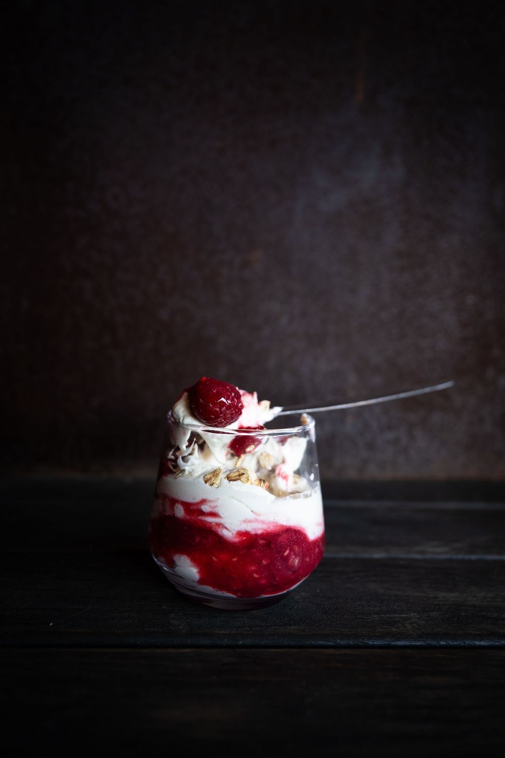 A layered dessert with red berry compote, whipped cream, and granola served in a glass jar against a dark background.