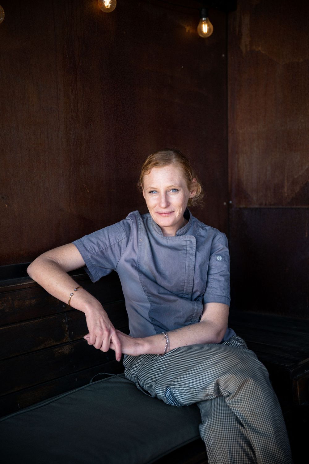 Chef in gray uniform poses casually against dark rustic wall with ambient lighting.