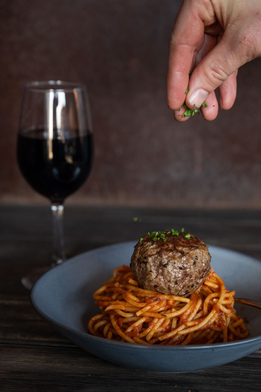 Plated dinner with pasta and meat being garnished, served alongside red wine in an elegant restaurant setting.