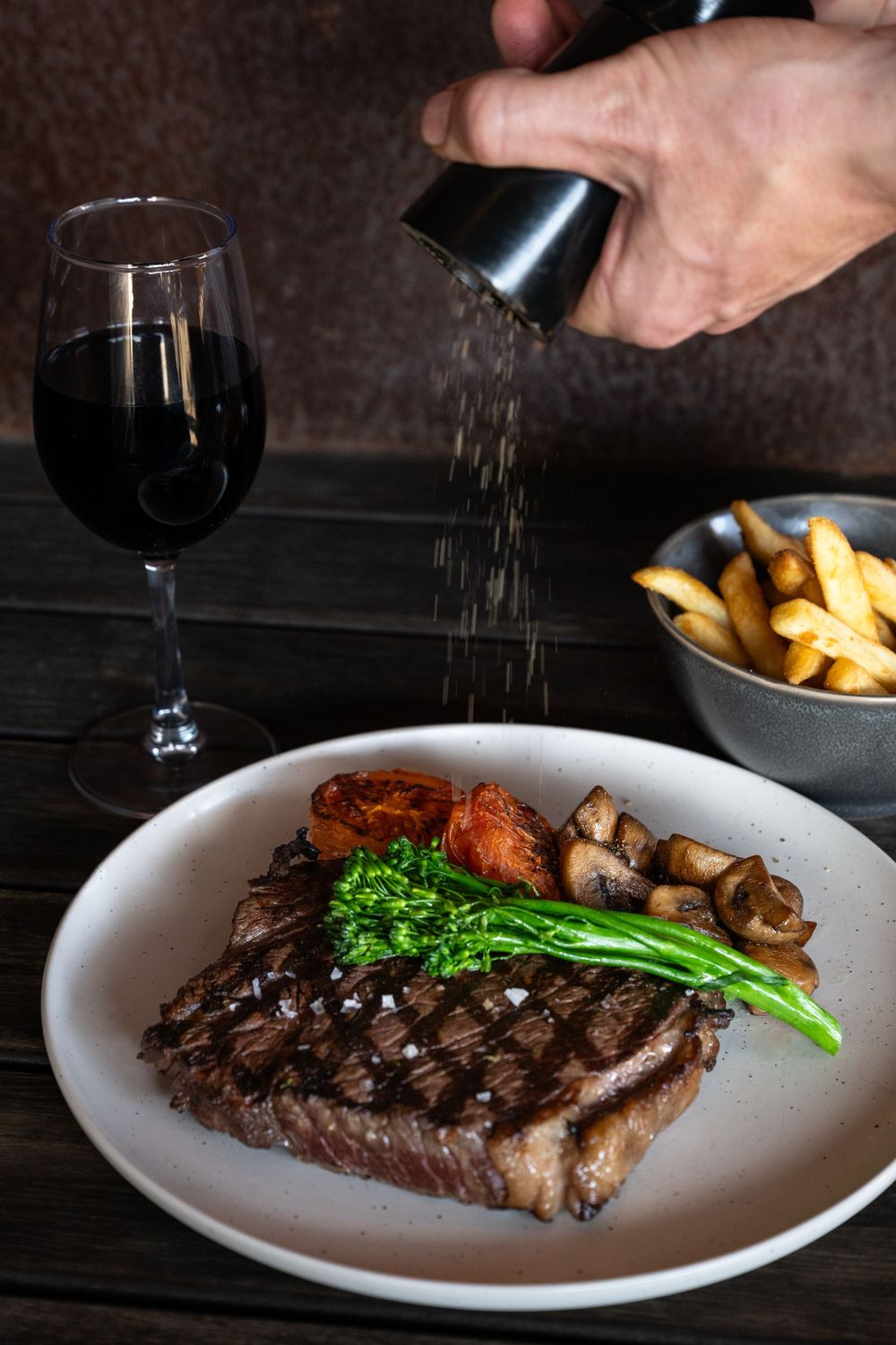 Grilled steak with broccolini and roasted tomatoes served alongside french fries and red wine on a rustic wooden table.