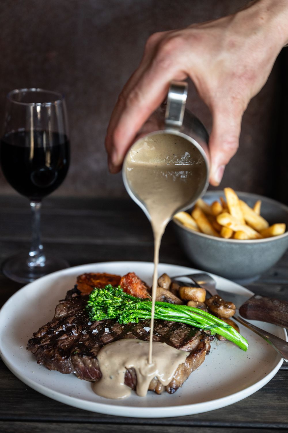 Someone pouring creamy peppercorn sauce over grilled steak with french fries and broccoli garnish on white plates.