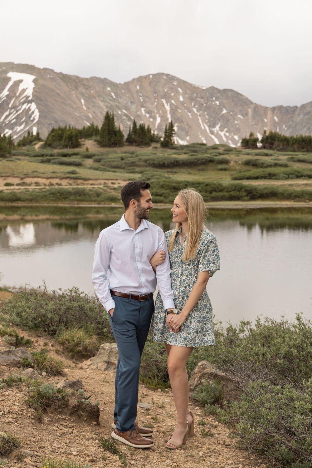 A couple stands together near an alpine lake with snow-capped mountains in the background on a cloudy day.