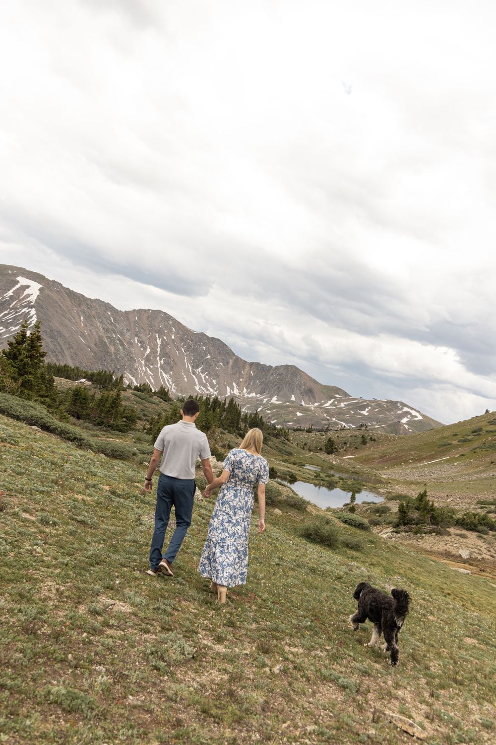 Two people and a dog walk along a scenic mountain trail with snow-capped peaks in the background on a cloudy day.