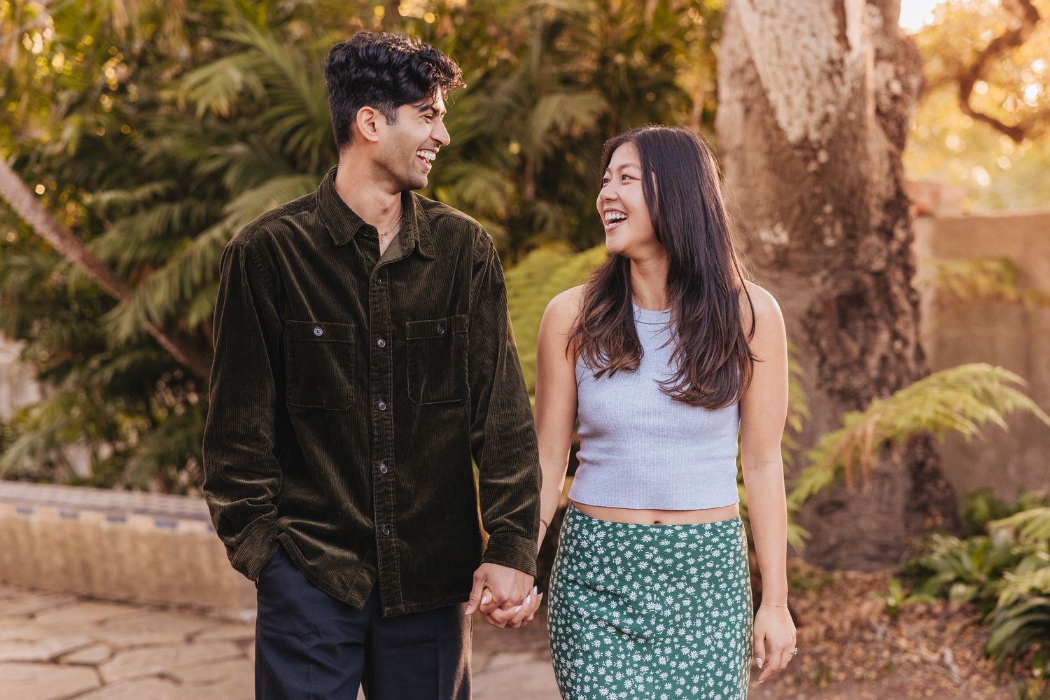 Young couple holding hands while walking together and laughing in a garden setting during golden hour.