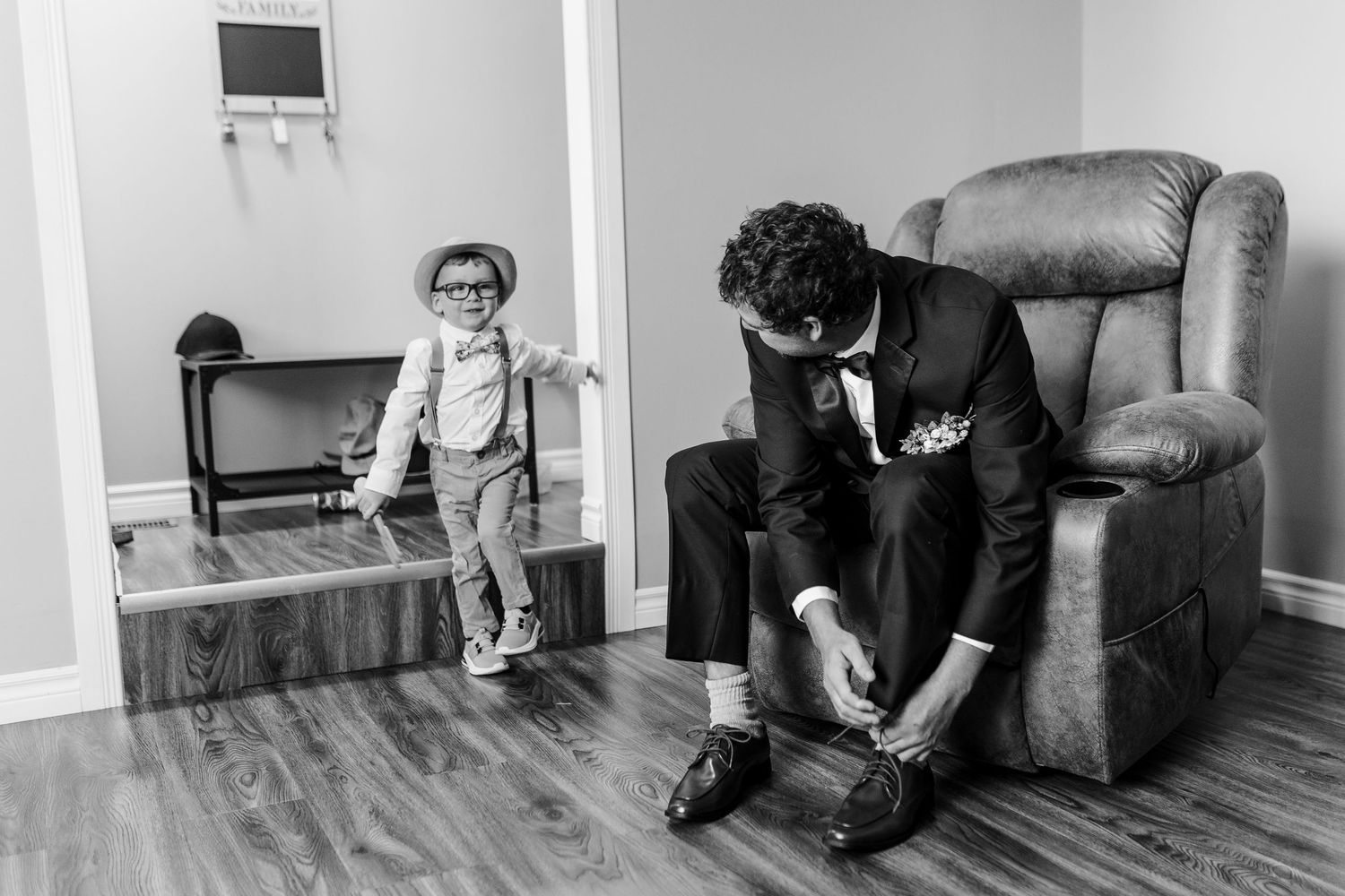 Black and white photograph showing two people getting ready in a room with a recliner chair and hardwood floors.