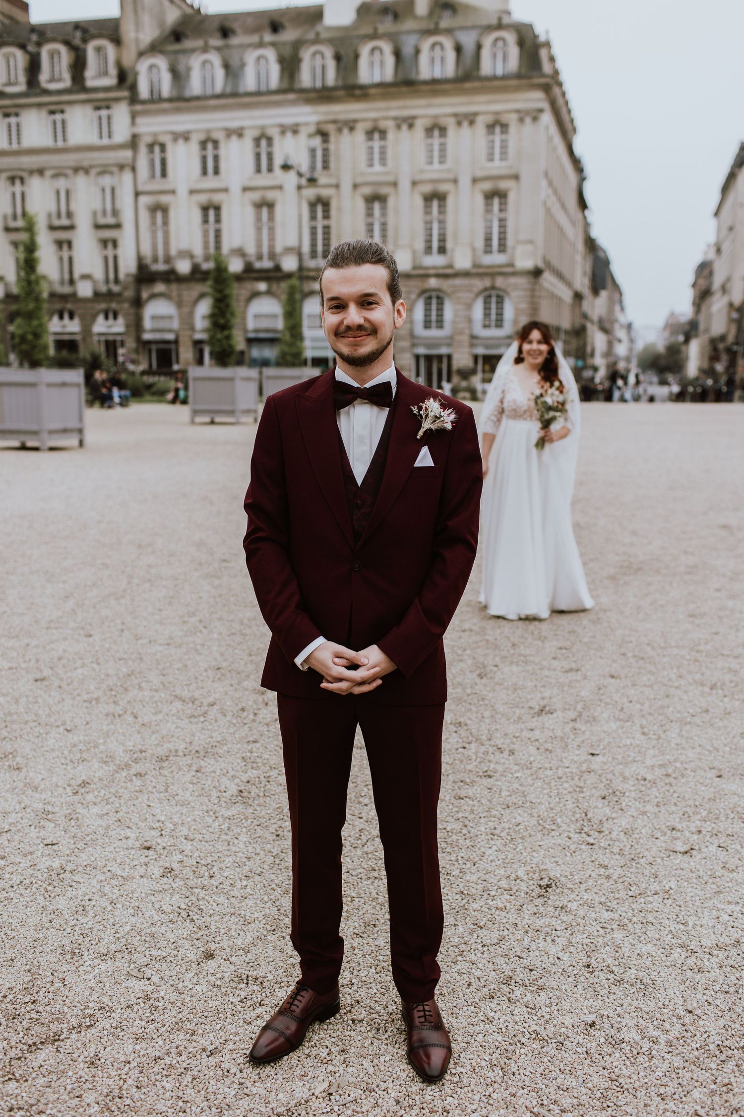 A wedding scene in front of an elegant European building with a groom in burgundy tuxedo and bride in white dress.