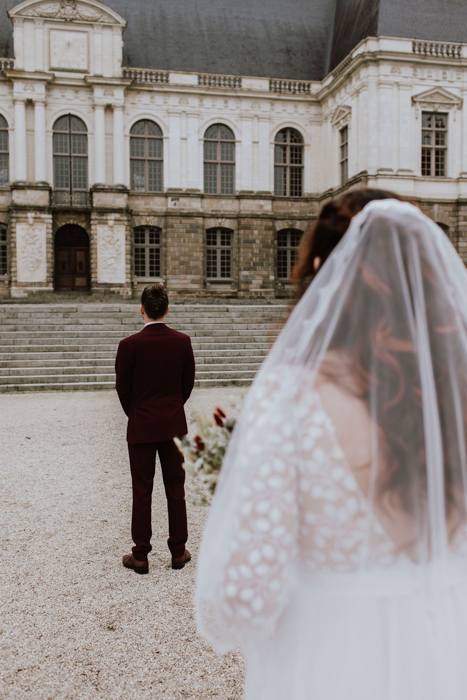 A couple stands in front of a historic white palace on their wedding day with the bride in white veil facing groom's back.