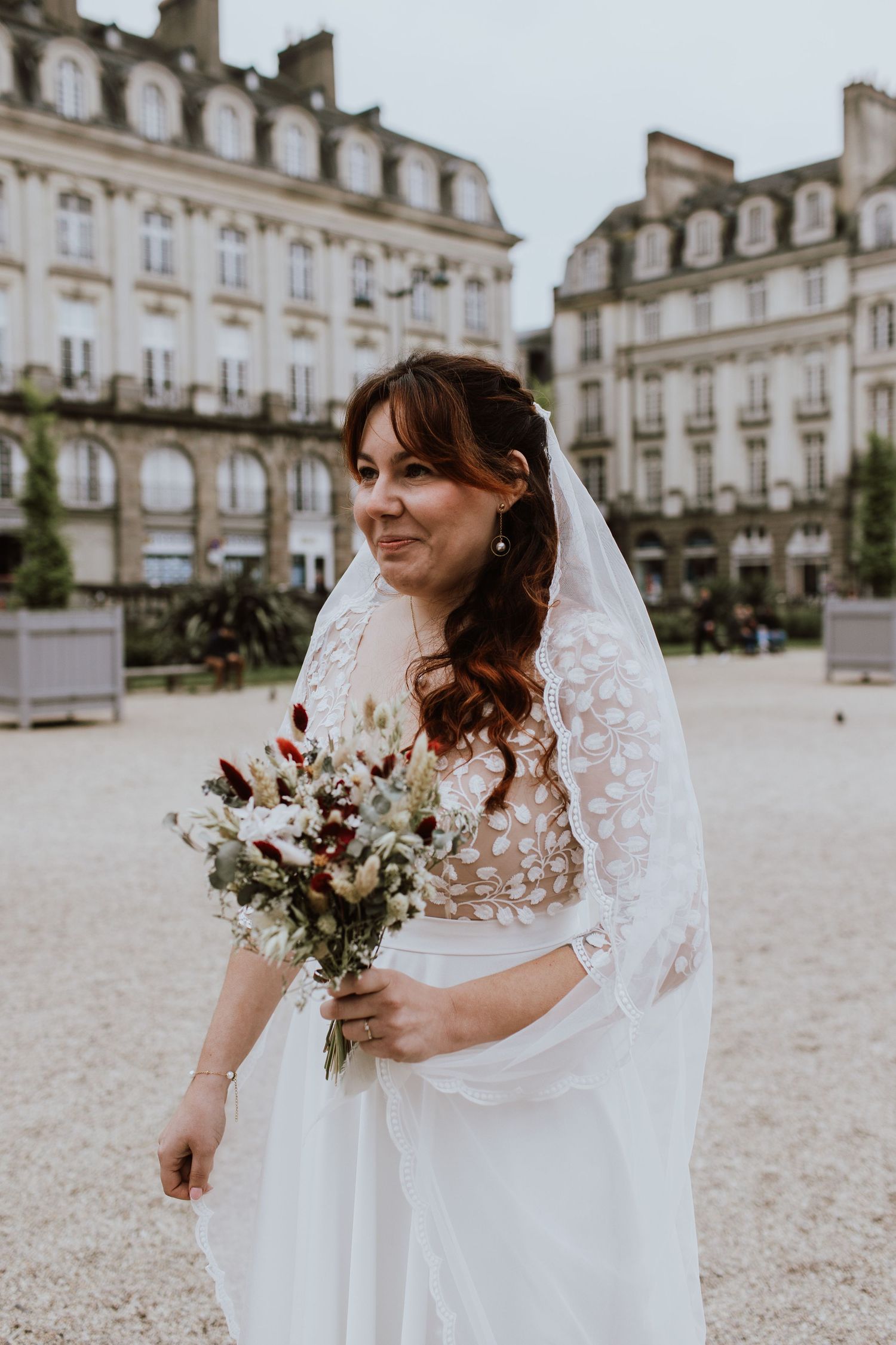 Bride in lace wedding dress holds a white and burgundy floral bouquet in front of a French chateau.