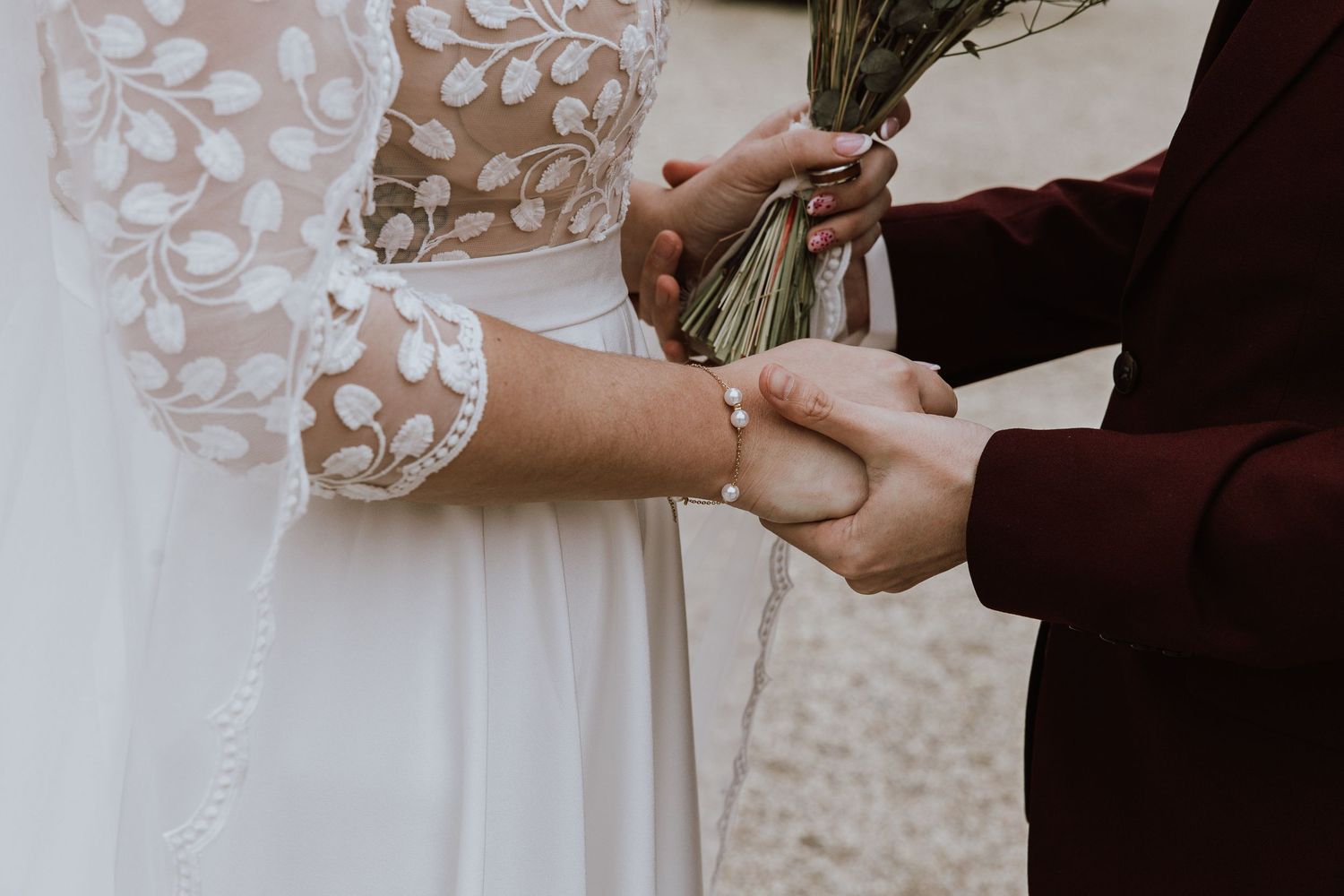 Close-up of bride and groom holding hands during wedding ceremony against white lace dress background.