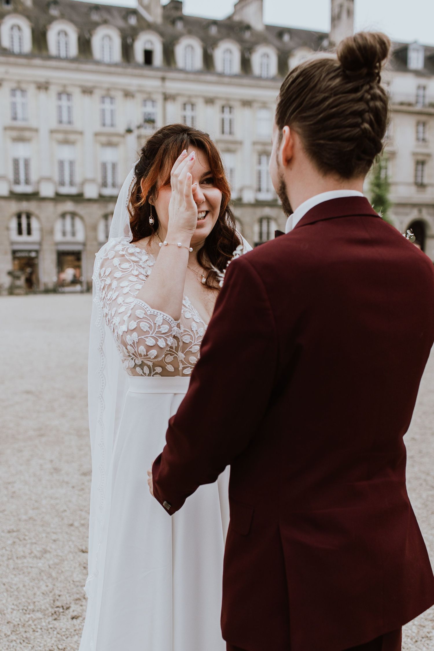 A couple in wedding attire share a candid moment outside an elegant European building with classic architecture.