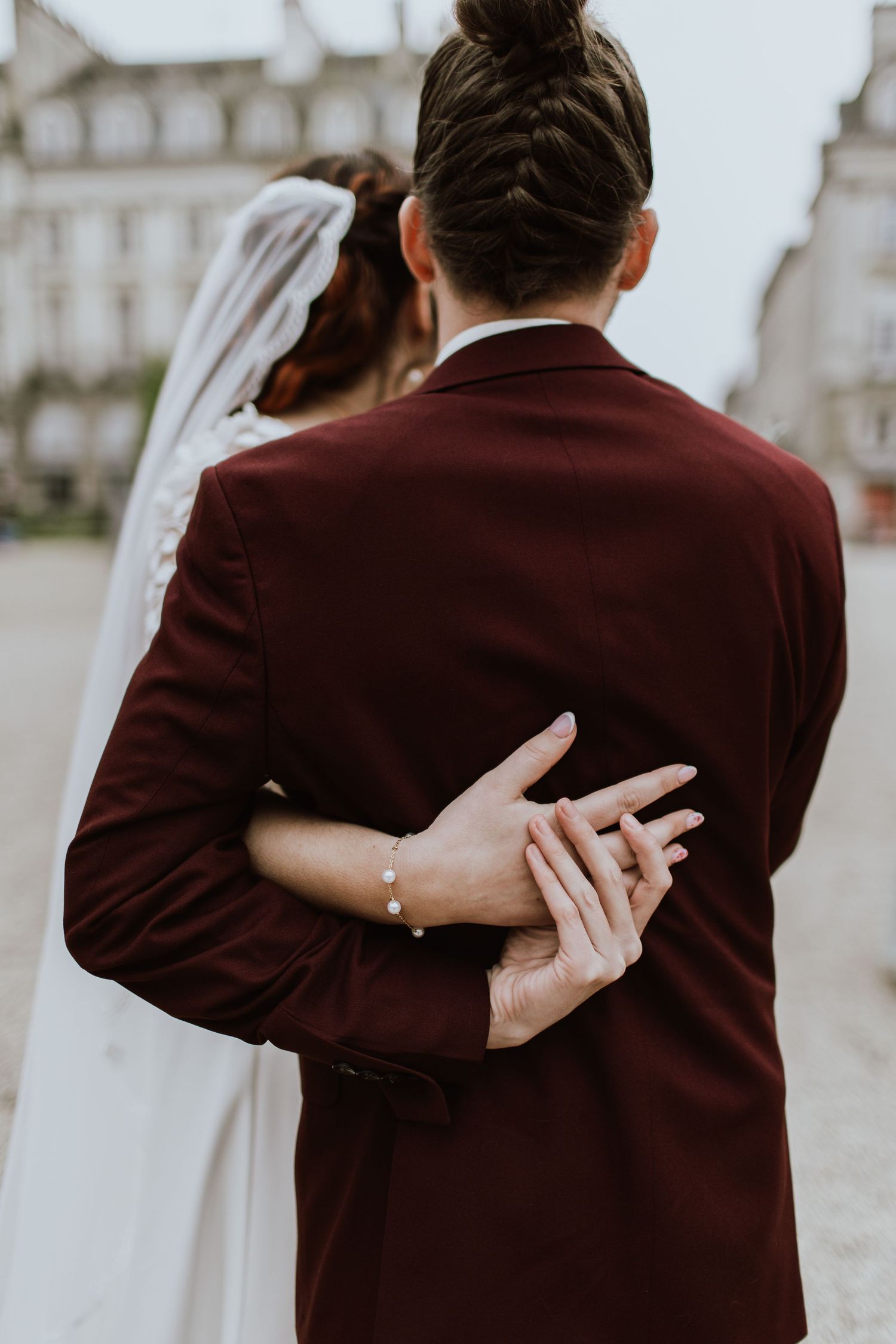 A romantic wedding photo showing couple embracing, with the bride's hand displaying ring and bracelet against groom's burgundy jacket.