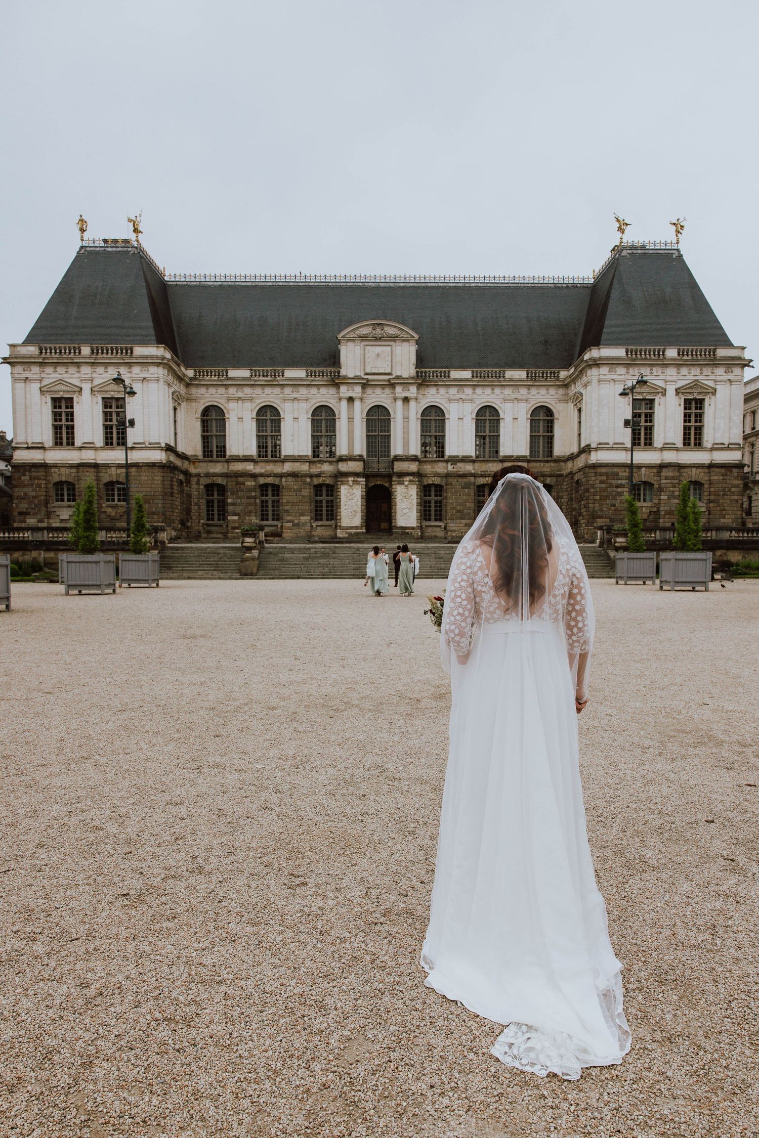 A bride in a flowing white gown and veil stands facing a grand French chateau with ornate architecture and towers.
