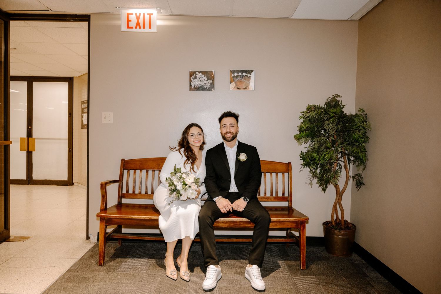 A couple in wedding attire sits on a wooden bench in a hallway near an exit sign and decorative plant.