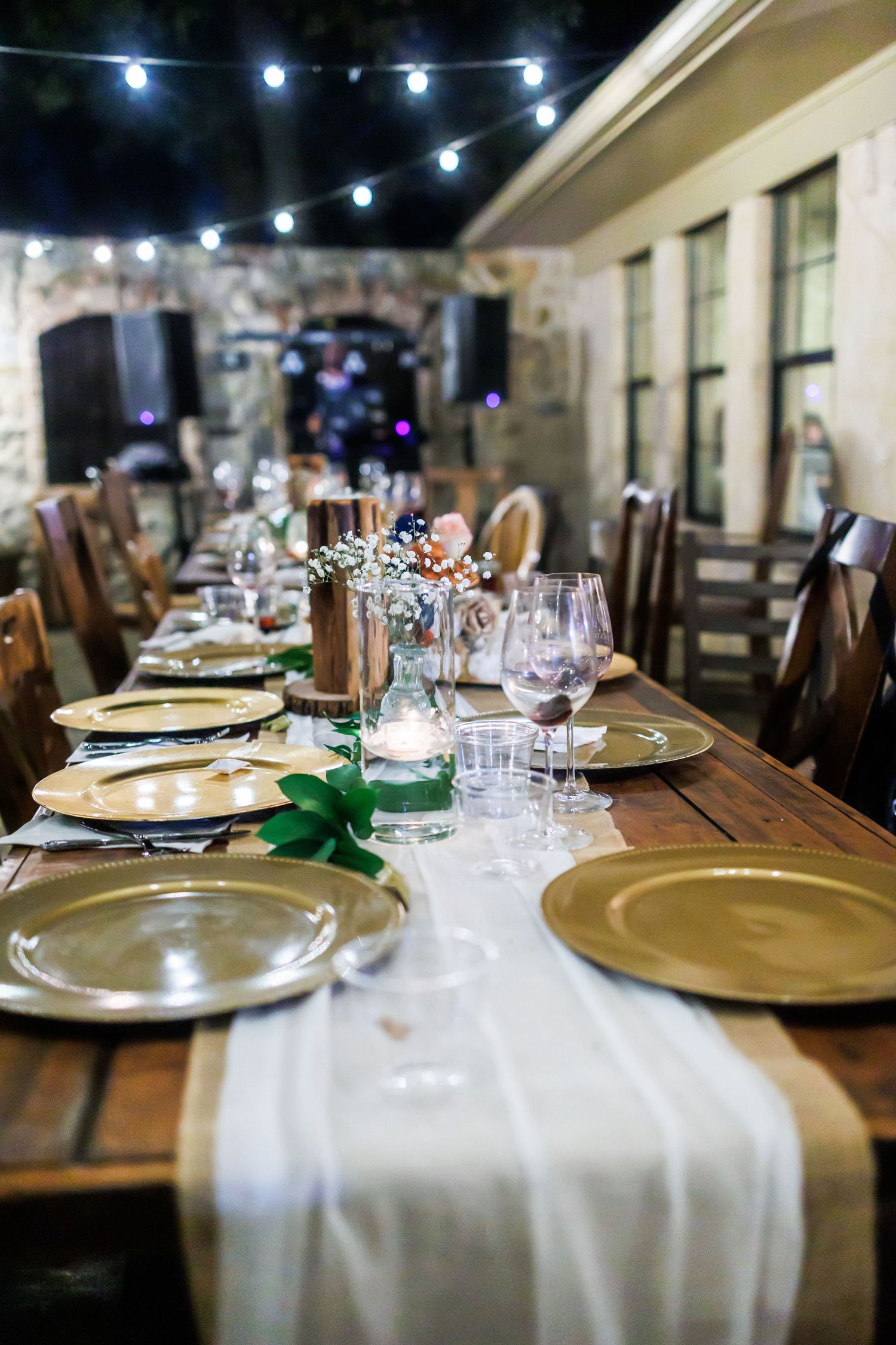 Elegant outdoor wedding table settings with gold charger plates and wine glasses under ambient string lighting.