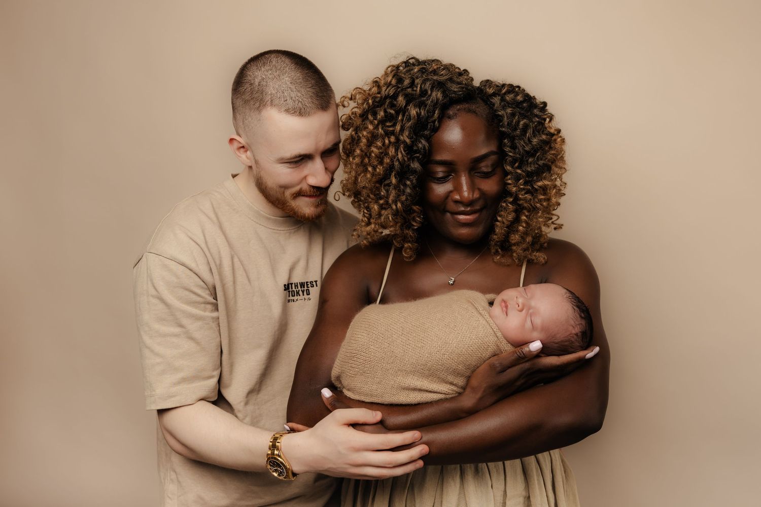 A couple cradles their newborn baby together against a neutral beige background.
