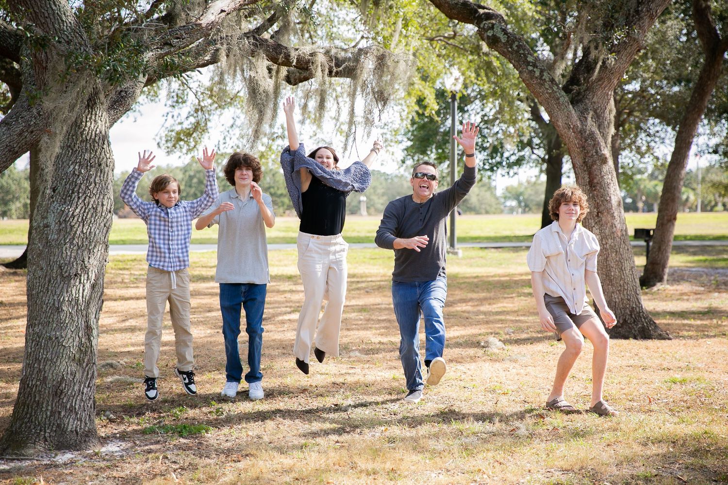 Group of people leaping and jumping together in a playful sequence on grass under large oak trees in a park setting.