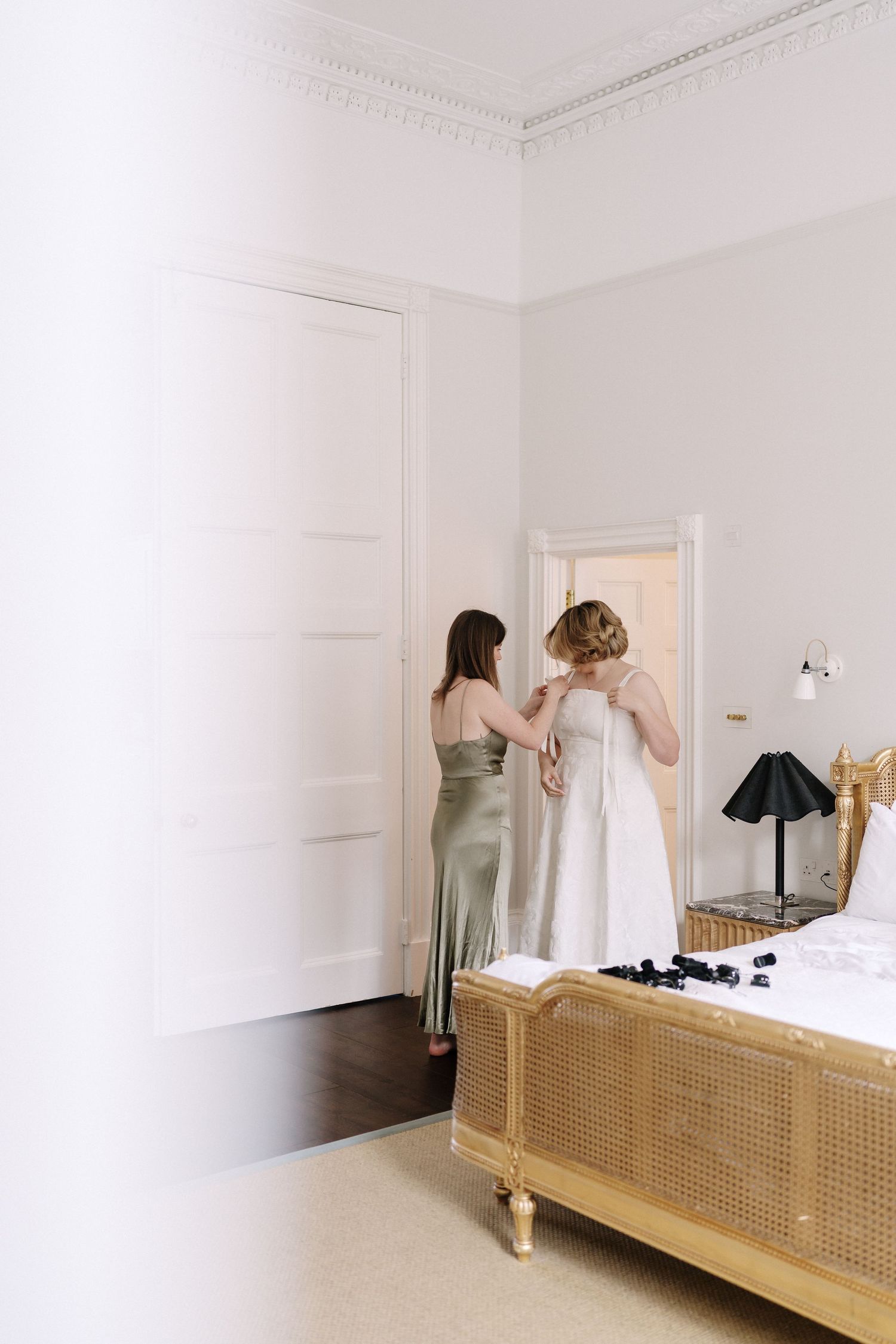 Two people getting dressed in an elegant white bedroom with a vintage-style rattan bed frame and table lamp.